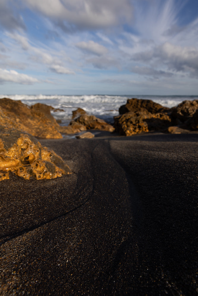 A low-angle shot of a black sand beach with wet, golden rocks in the foreground. The ocean waves crash in the background under a cloudy sky with streaks of white clouds.