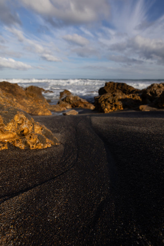 A low-angle shot of a black sand beach with wet, golden rocks in the foreground. The ocean waves crash in the background under a cloudy sky with streaks of white clouds.