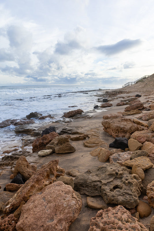 A rocky beach with waves crashing on the shore under a cloudy sky. A wooden staircase leads up a sandy dune on the right side of the image.