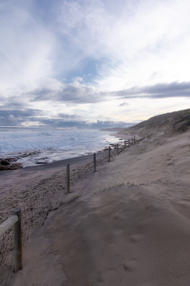 A sandy beach with a wire fence along a dune. The ocean waves crash on the shore under a cloudy sky.