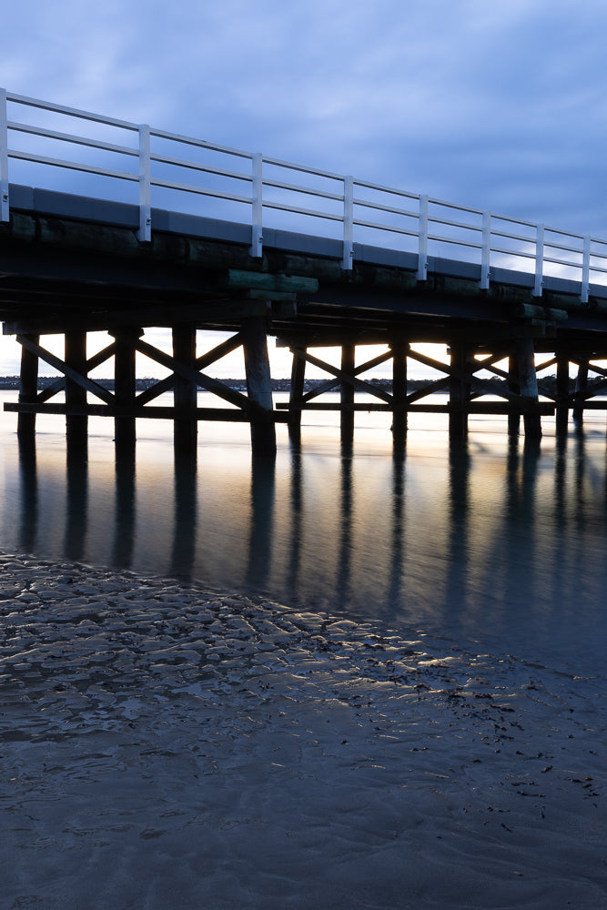 Pier pilings cast long shadows on calm water at dusk.