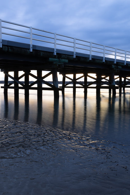 Pier pilings cast long shadows on calm water at dusk.