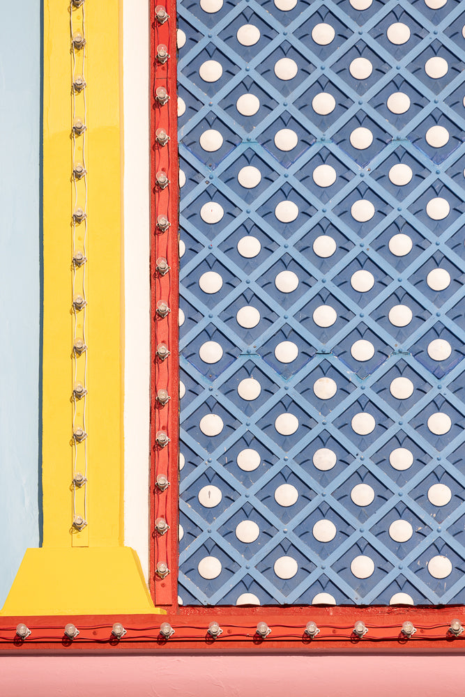 A close-up of a colorful, textured wall with a blue diamond pattern and white circles, bordered by red trim with exposed light bulbs. A yellow vertical beam with a string of lights is on the left.