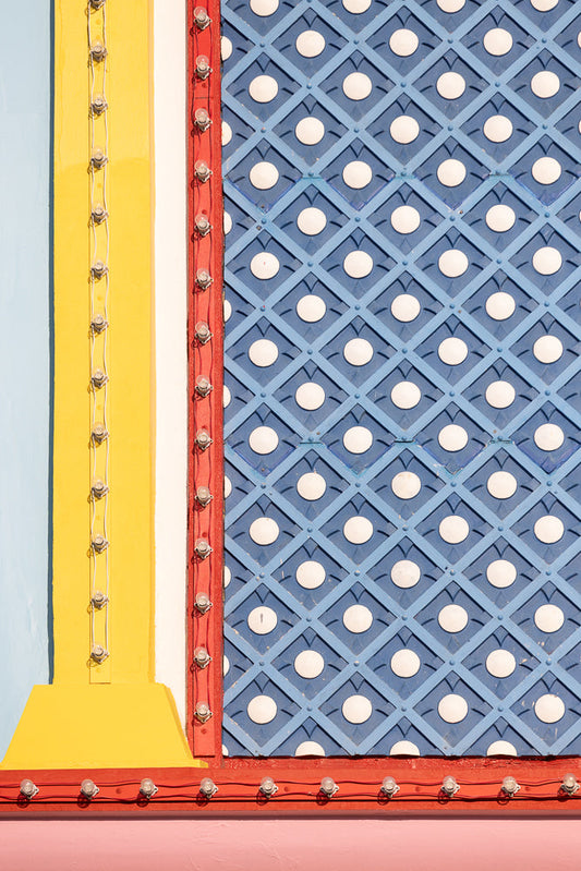 A close-up of a colorful, textured wall with a blue diamond pattern and white circles, bordered by red trim with exposed light bulbs. A yellow vertical beam with a string of lights is on the left.