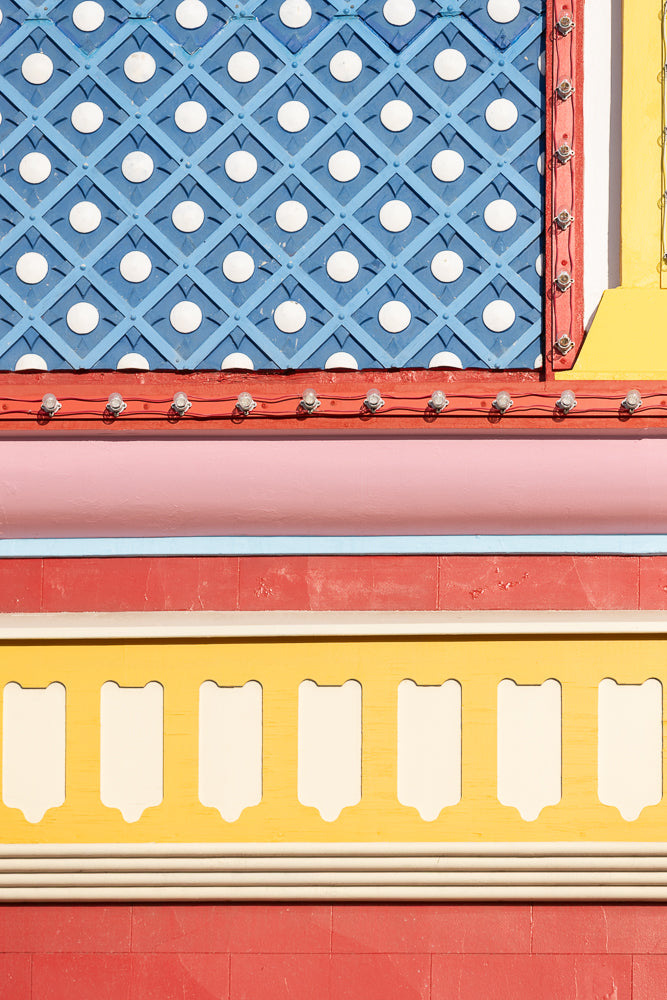 A close-up view of a colorful building facade. The top section features a blue diamond pattern with white circles. Below this is a red band with a string of clear light bulbs. Further down, there is a pink band, a thin blue band, and a red band. The bottom section shows a yellow band with white decorative elements and a red brick pattern.