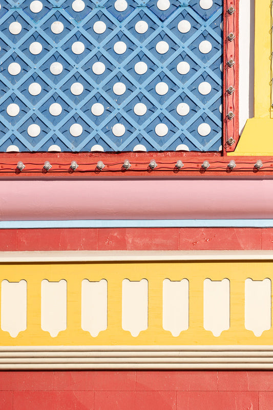 A close-up view of a colorful building facade. The top section features a blue diamond pattern with white circles. Below this is a red band with a string of clear light bulbs. Further down, there is a pink band, a thin blue band, and a red band. The bottom section shows a yellow band with white decorative elements and a red brick pattern.