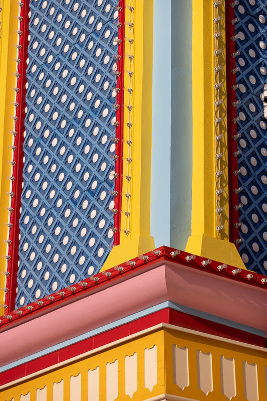A close-up shot of a brightly colored building facade with a blue diamond pattern and white circles, trimmed with red and yellow. Strings of clear light bulbs run along the edges of the yellow and red trim.