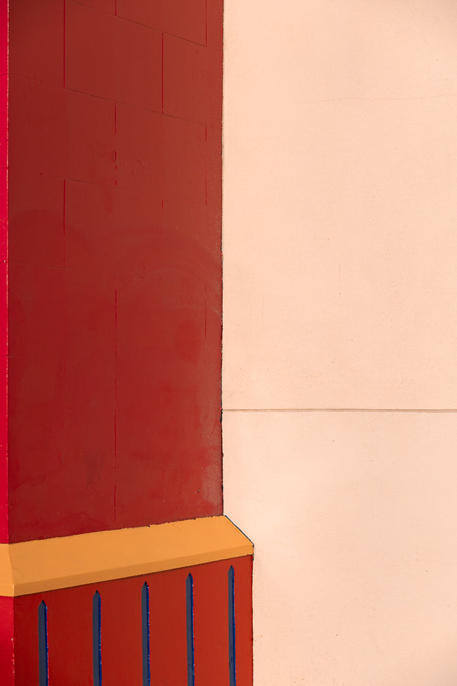 A close-up abstract shot of a building corner. A dark red wall with vertical blue lines and a yellow trim meets a light peach-colored wall.