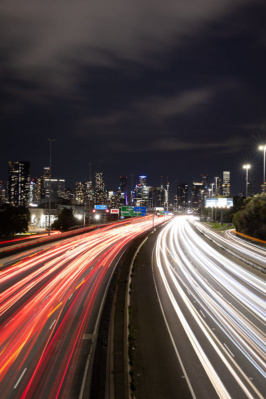 A long exposure shot of a highway at night, with streaks of red and white light from car headlights and taillights creating a dynamic effect. The city skyline is visible in the background under a cloudy sky.