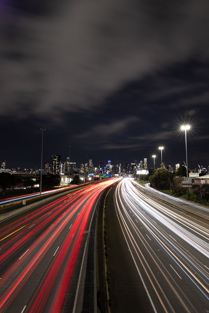 A long exposure shot of a highway at night, with streaks of red and white light from car headlights and taillights creating a dynamic, abstract pattern against the dark sky and city skyline in the distance.