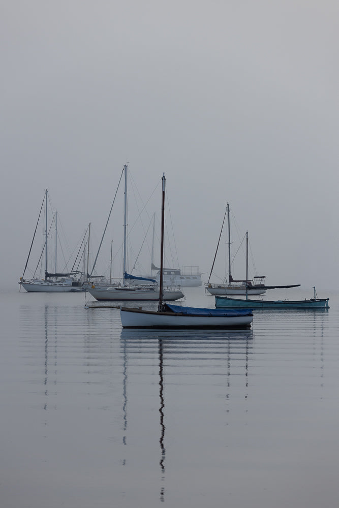 Several sailboats and a small rowboat are moored on a calm body of water under a hazy, overcast sky. The boats are reflected in the still water, creating a serene and peaceful atmosphere.