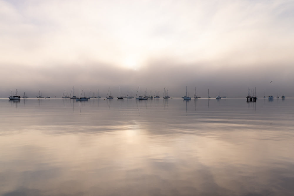 A fleet of sailboats and motorboats are anchored on a calm body of water under a hazy, overcast sky. The sun is diffused by the clouds, casting a soft glow on the scene. The reflections of the boats and sky are visible on the water's surface.