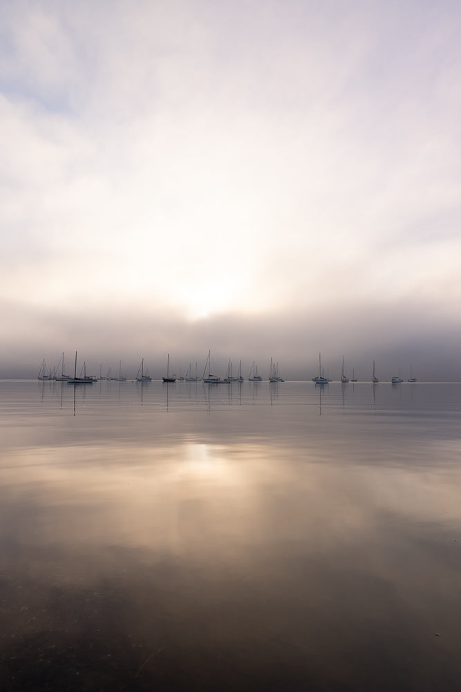 A fleet of sailboats are anchored on a calm body of water under a hazy, overcast sky. The sun is diffused by the clouds, casting a soft glow on the scene and reflecting on the water's surface.