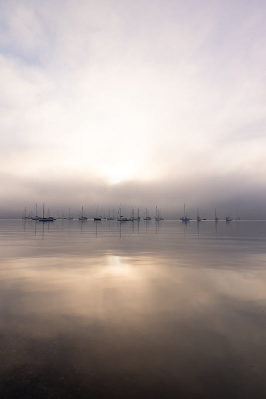 A fleet of sailboats are anchored on a calm body of water under a hazy, overcast sky. The sun is diffused by the clouds, casting a soft glow on the scene and reflecting on the water's surface.