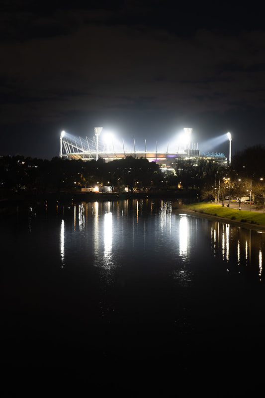 A stadium is illuminated by bright lights at night, with its reflection shimmering on the dark water of a river in the foreground. Trees and streetlights line the riverbank.