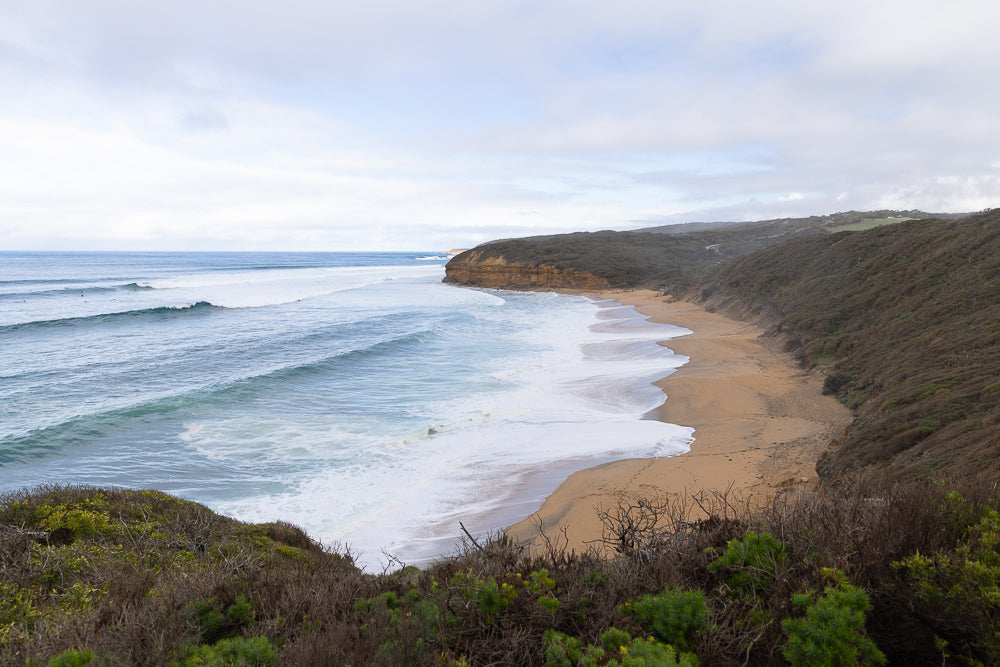A wide, sandy beach curves along the coast, with gentle waves rolling onto the shore. Cliffs rise on the right, covered in dense, dry vegetation. The ocean stretches to the horizon under a cloudy sky.