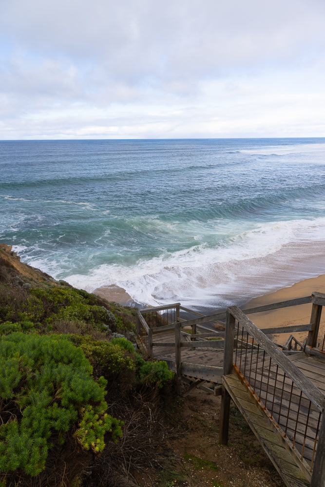 A wooden staircase descends towards a sandy beach where waves crash onto the shore under a cloudy sky.