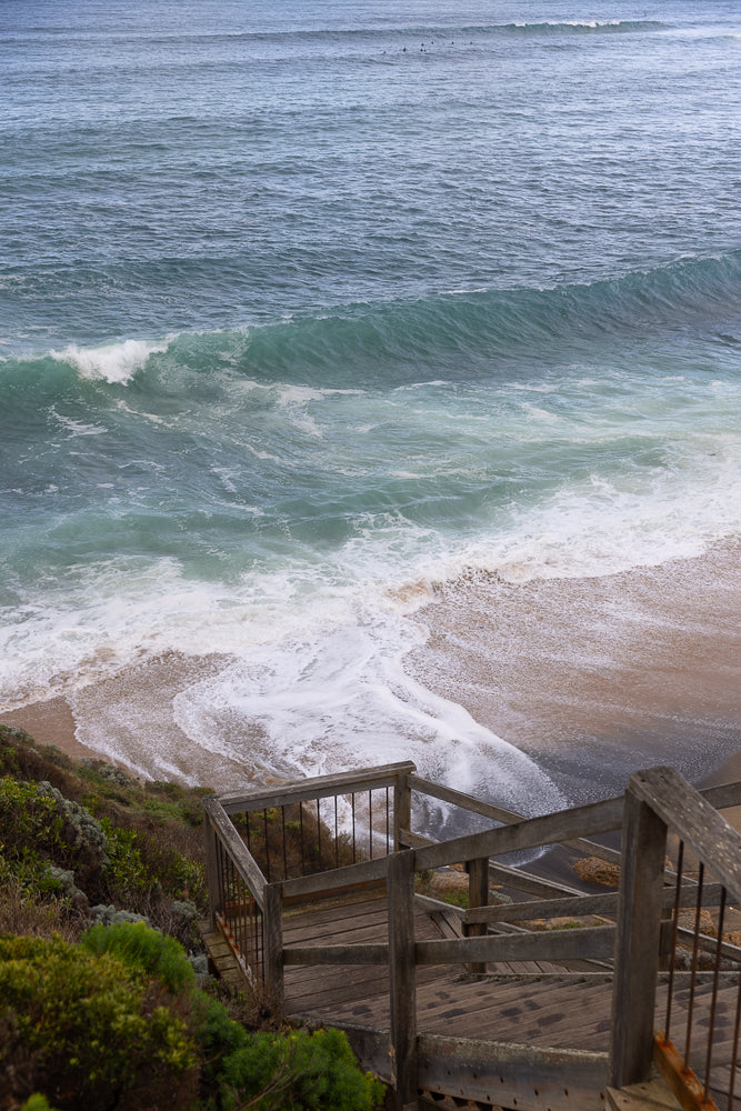 A wooden staircase with metal railings leads down to a sandy beach where waves are crashing onto the shore. The ocean is a deep blue-green with white foam from the waves.