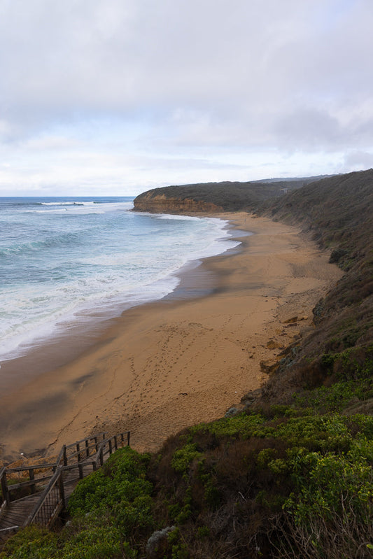 A wide view of a sandy beach with gentle waves rolling onto the shore under a cloudy sky. A wooden staircase leads down to the beach from a grassy cliffside.