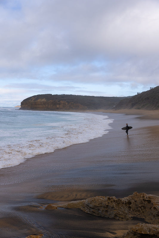 A lone surfer walks along a wet sandy beach carrying a surfboard. Waves crash on the shore, and a large cliff face covered in vegetation rises in the background under a cloudy sky.
