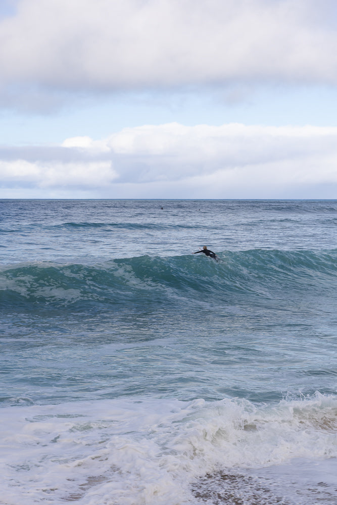 A surfer rides a wave on a cloudy day. The surfer is silhouetted against the bright white foam of the wave. The ocean is a deep blue, and the sky is filled with white clouds.