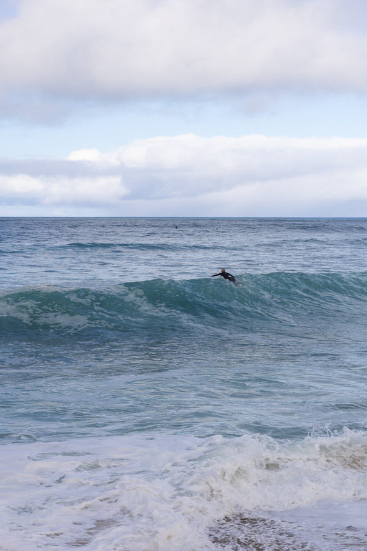 A surfer rides a wave on a cloudy day. The surfer is silhouetted against the bright white foam of the wave. The ocean is a deep blue, and the sky is filled with white clouds.