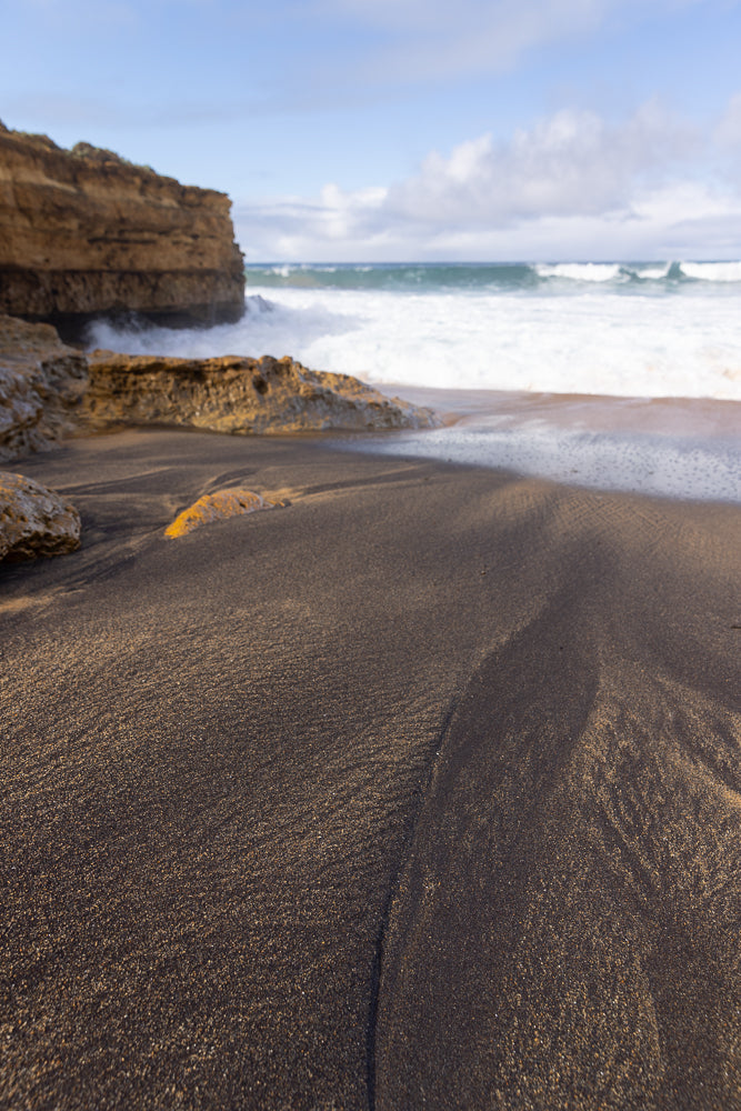 A low-angle shot of dark, wet sand on a beach, with ripples and patterns from receding waves. In the background, rough, tan-colored rock formations meet the ocean, where white waves crash against the shore under a blue sky with scattered clouds.