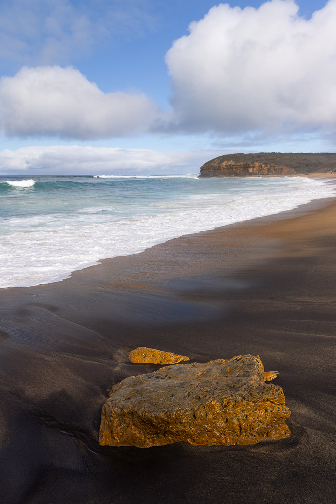 A wide shot of a beach with dark sand and a large, wet, golden rock in the foreground. The ocean has turquoise water with white waves crashing onto the shore. In the background, a cliff face covered in vegetation rises from the sea under a blue sky with fluffy white clouds.