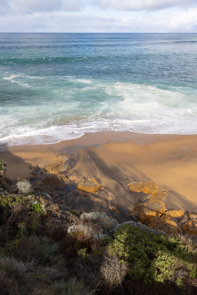 A scenic view of a beach with waves crashing onto the shore. The sand has dark streaks, and rocks are scattered along the waterline. Lush green and grey vegetation covers the foreground.