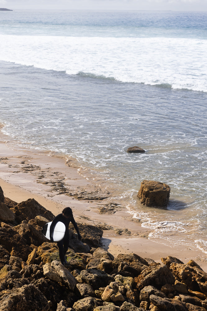A surfer in a black wetsuit walks over rocky terrain towards the ocean, carrying a white surfboard.