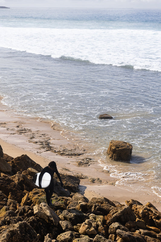 A surfer in a black wetsuit walks over rocky terrain towards the ocean, carrying a white surfboard.