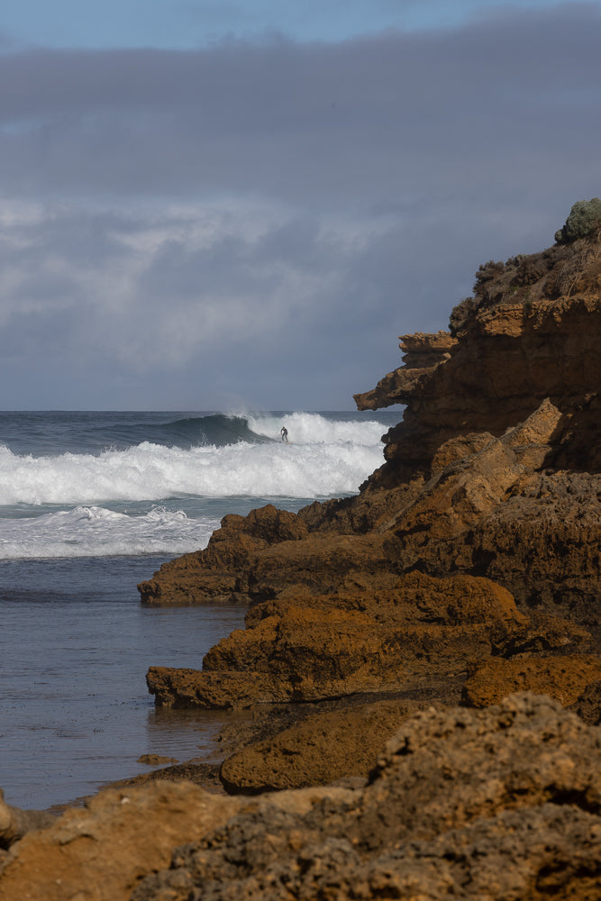A surfer rides a wave near a rocky coastline under a cloudy sky.