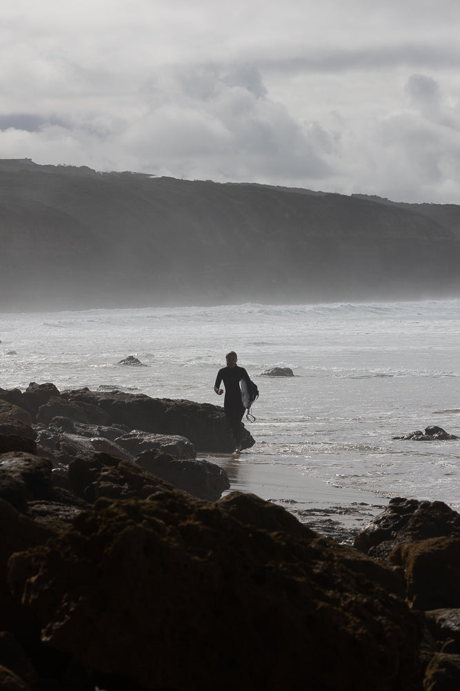 A surfer in a black wetsuit walks out of the ocean carrying a surfboard. They are walking on a rocky shore with waves crashing in the background. A misty, cloudy sky hangs over a distant, dark, tree-covered hill.