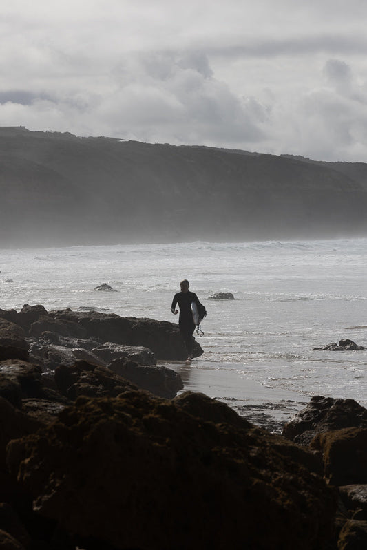 A surfer in a black wetsuit walks out of the ocean carrying a surfboard. They are walking on a rocky shore with waves crashing in the background. A misty, cloudy sky hangs over a distant, dark, tree-covered hill.