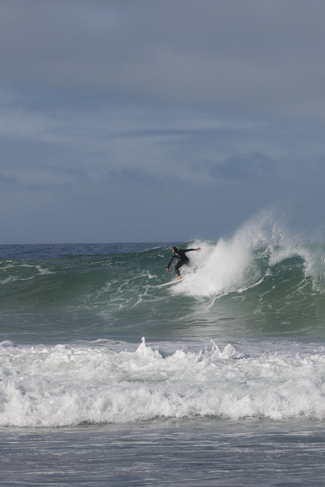 A surfer in a black wetsuit rides a wave, carving a turn as white spray erupts around them. The wave is green and translucent, with white foam cresting at the top. The ocean is a deep blue under a cloudy sky.
