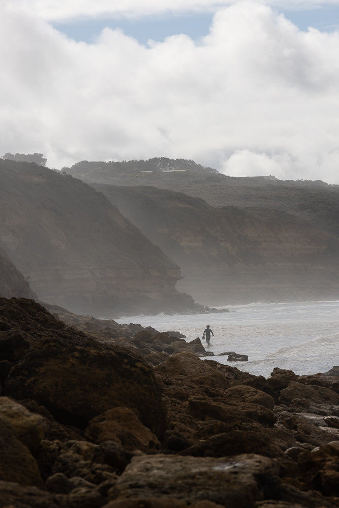 A lone surfer walks into the ocean on a rocky shore, with misty cliffs and cloudy skies in the background.