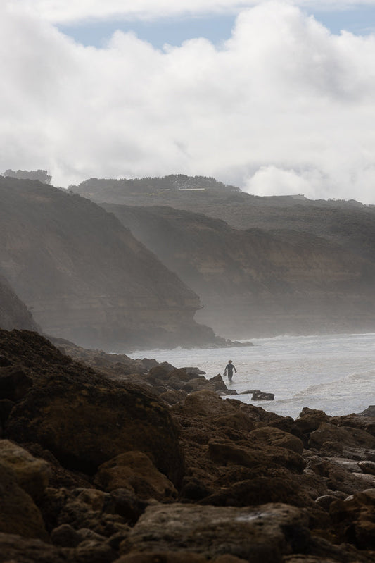 A lone surfer walks into the ocean on a rocky shore, with misty cliffs and cloudy skies in the background.