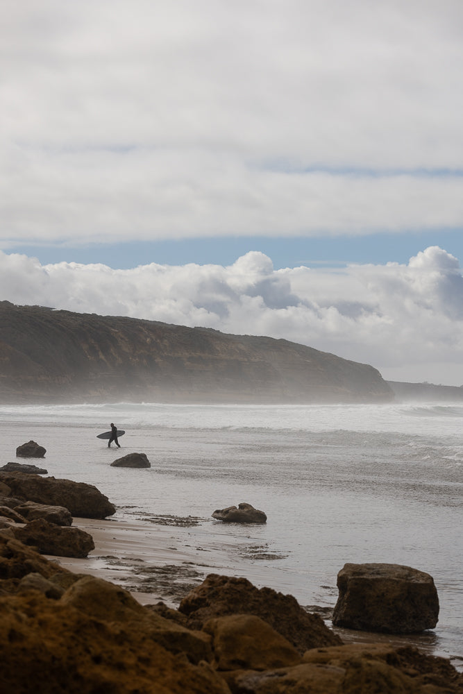 A lone surfer walks into the ocean carrying a surfboard, with waves crashing against a rocky coastline in the background.