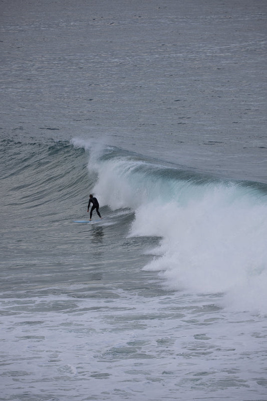 A surfer in a black wetsuit rides a wave on a light blue surfboard. The surfer is positioned on the left side of the frame, facing right, as the wave curls and breaks to the right.