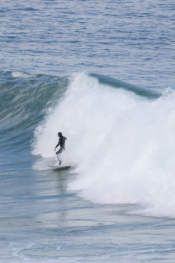 A surfer in a black wetsuit rides a wave, carving through the water as a large plume of white spray erupts behind them.