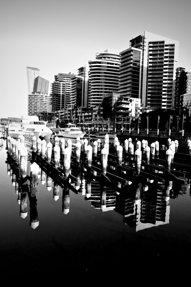 A black and white image shows a marina with several boats docked. The foreground is dominated by a row of wooden posts with white caps, reflected in the calm water. In the background, a cityscape of modern buildings rises against a clear sky. The reflections of the buildings and posts create a symmetrical and artistic composition.