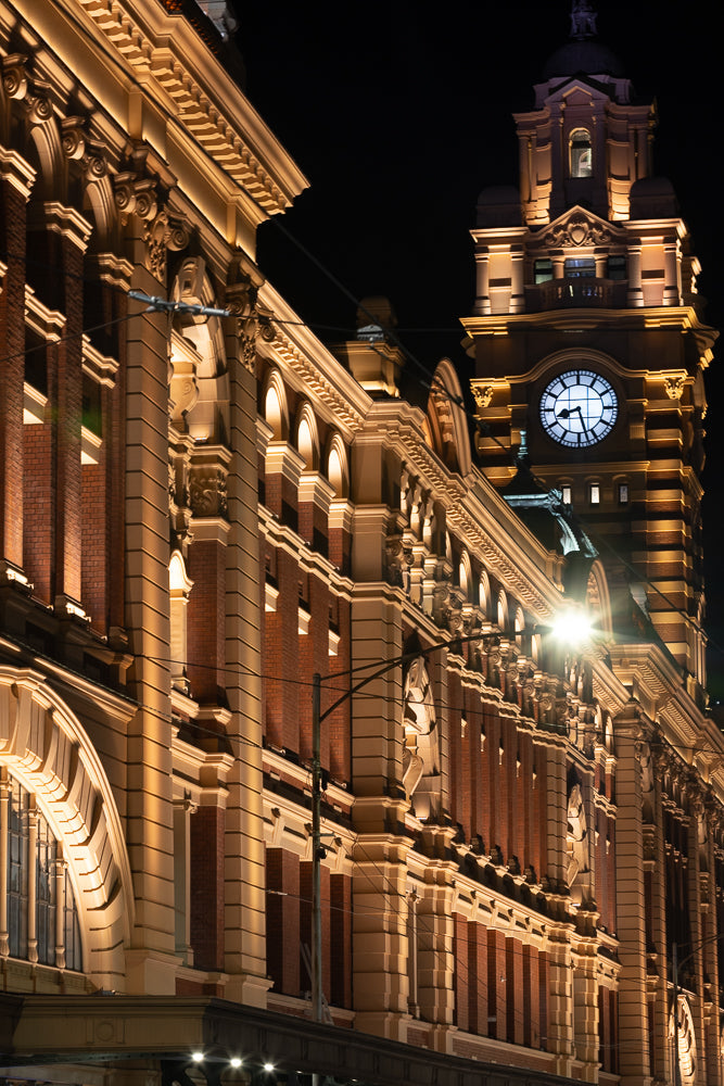 A grand, ornate building with a prominent clock tower is illuminated at night. The architecture features brickwork, decorative stonework, and arched windows, all highlighted by warm, golden lighting.