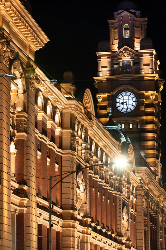 A close-up, low-angle night shot of the ornate facade of a historic building, illuminated by warm yellow lights. The image focuses on the detailed architecture, including brickwork, decorative cornices, arched windows, and a prominent clock tower with a large, illuminated clock face.