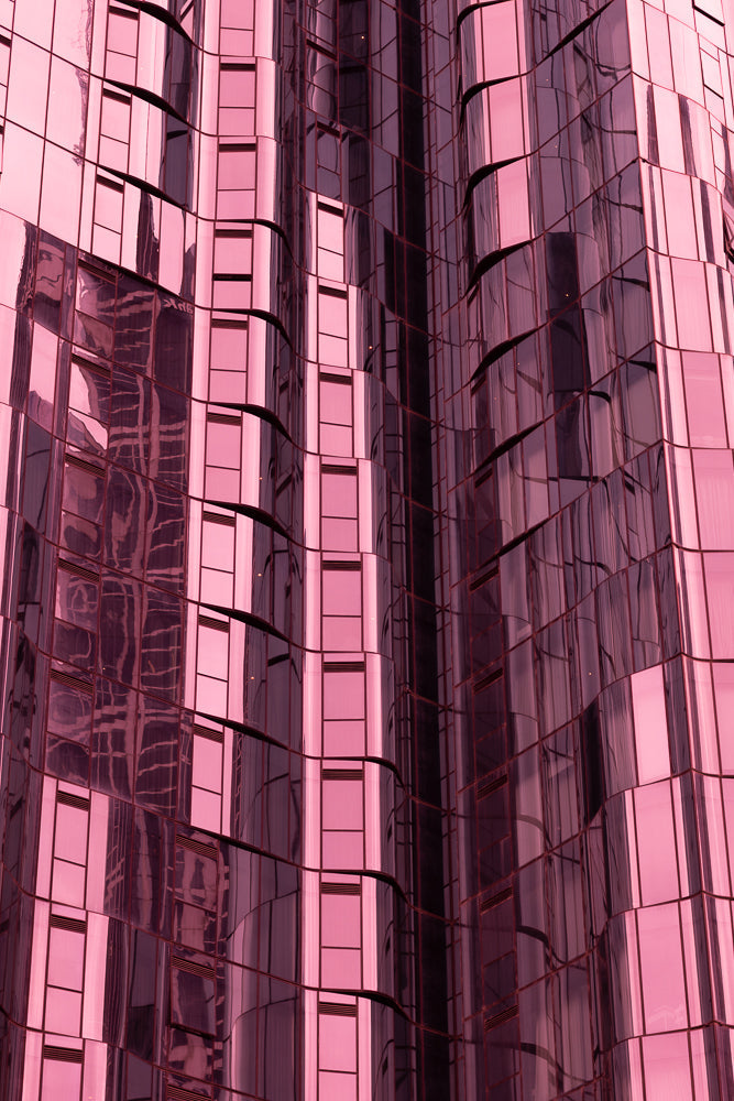 A close-up, abstract view of a modern building facade with pink-tinted glass panels. The glass reflects the surrounding environment, creating distorted patterns and lines. The building's structure appears to curve and undulate, giving it a dynamic and fluid appearance.
