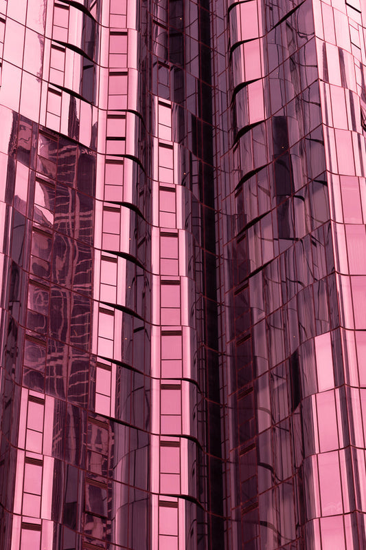 A close-up, abstract view of a modern building facade with pink-tinted glass panels. The glass reflects the surrounding environment, creating distorted patterns and lines. The building's structure appears to curve and undulate, giving it a dynamic and fluid appearance.