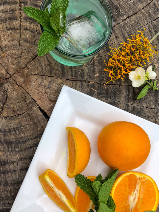 A refreshing drink with a large ice cube and mint leaves sits on a rustic wooden surface next to a white plate of sliced oranges and more mint.