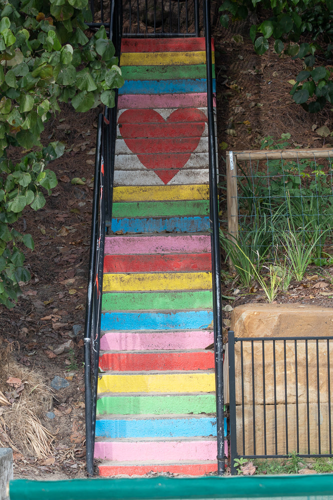 A set of outdoor stairs painted with rainbow colors, featuring a large red heart painted on the top steps. The stairs are bordered by black railings and surrounded by greenery and natural ground cover.
