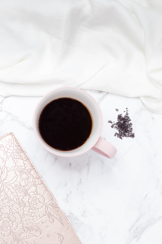 A pink mug filled with black coffee sits on a marble surface next to a small pile of dark crystals and a light pink notebook with embossed floral patterns. A white cloth is draped behind the mug.