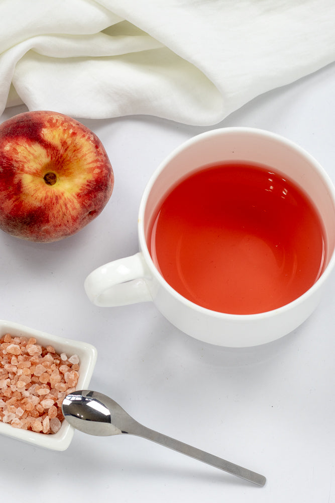 A ripe peach sits next to a white mug filled with a red beverage. A small white dish contains pink Himalayan salt, and a silver spoon rests nearby on a white surface, with a white cloth draped in the background.