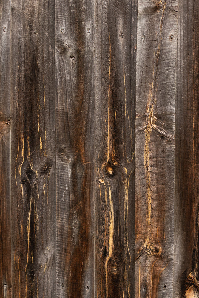 A close-up, vertical shot of weathered, dark brown wooden planks. The planks have visible grain patterns, knots, and some lighter brown or golden streaks. Several nails are visible, securing the planks.